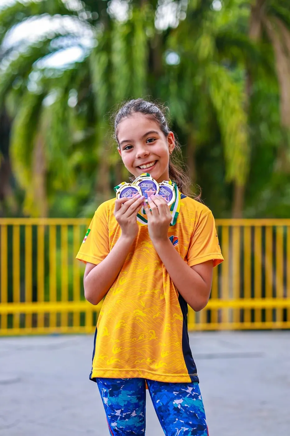 Lelê sorrindo ao ar livre com várias medalhas nas mãos